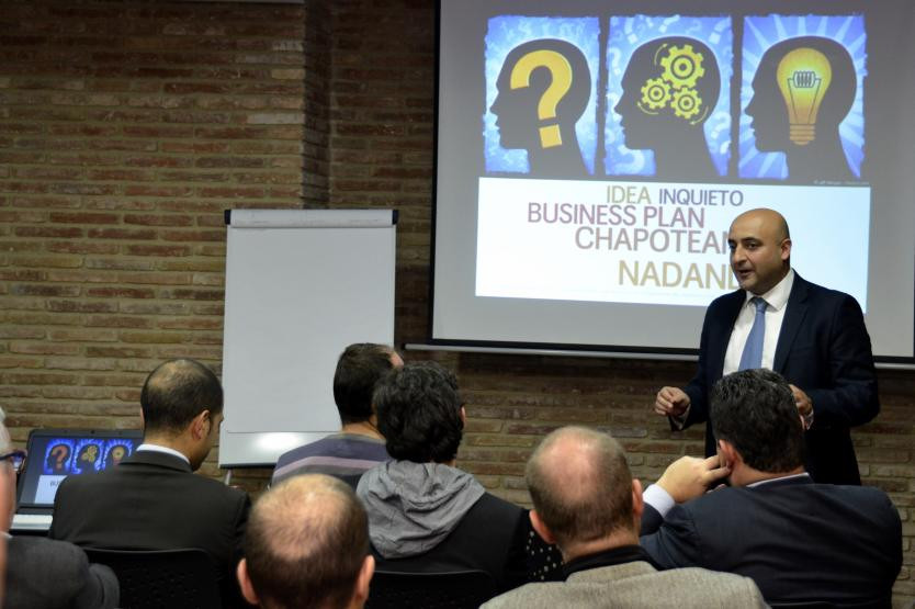 Presentador en una sala de conferencias explicando un plan de negocio frente a una audiencia, con una proyección de imágenes de perfiles humanos y conceptos de innovación.